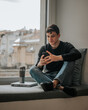 © ADDICTIVE STOCK - Smiling teenager browsing smartphone on windowsill