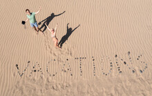 Overhead View Of A Man And Woman Standing In Sand Dunes Next To The Word Vacations, Maspalomas, Gran Canaria, Canary Islands, Spain