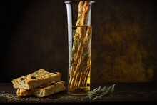  A Glass Of Water And Some Bread Sticks In A Vase And A Bottle Of Oil And Some Sage Leaves On A Table With A Black Background Of Brown Paper And Gold Cloth With A Black.