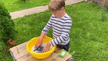 The Boy Helps His Mother In The Fresh Air. Washes Socks With Soap And Hands In A Yellow Basin.