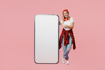 Wall Mural - Full body fun young woman wear white t-shirt red hat point on big huge blank screen mobile cell phone smartphone with workspace copy space mockup area isolated on plain pastel light pink background.