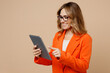 © ViDi Studio - Side view young fun employee business woman corporate lawyer 30s wear classic formal orange suit glasses work in office hold use digital tablet pc computer isolated on plain beige background studio.
