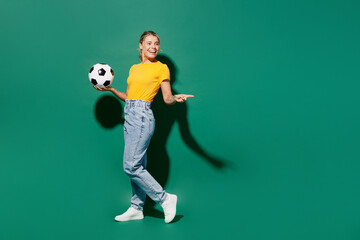 Wall Mural - Full body fun young woman fan wear yellow t-shirt cheer up support football sport team hold in hand soccer ball watch tv live stream point index finger aside on area isolated on dark green background.