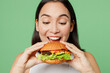 Close up young happy cheerful woman wear white clothes holding eating biting tasty burger isolated on plain pastel light green background. Proper nutrition healthy fast food unhealthy choice concept.
