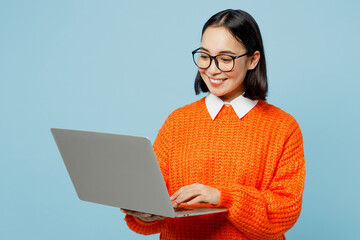 Young smiling fun smart IT woman of Asian ethnicity wear orange sweater glasses hold use work on laptop pc computer isolated on plain pastel light blue cyan background studio People lifestyle concept