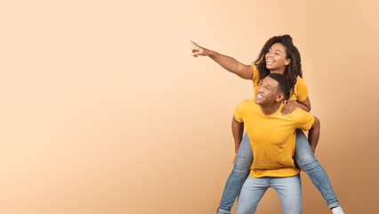 Excited black couple having fun, lady piggybacking guy and pointing at copy space, yellow background, panorama