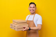 © Bangun Stock Photo - Smiling young Asian man barista barman employee wear brown apron working in coffee shop holding italian pizza in cardboard flat box looking camera on yellow background. Small business startup concept