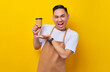 © Bangun Stock Photo - Excited young Asian man barista barman employee wearing brown apron work in coffee shop showing paper cup of coffee or tea on hand isolated on yellow background. Small business startup