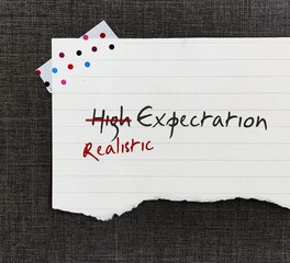 Stick note with handwritten HIGH EXPECTATION crossed HIGH off, replaced with REALISTIC, to be able to move forward in life with more acceptance when things turn out lower than expected.