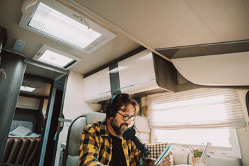 Wall Mural - One modern traveler young adult man speaking at the microphone in podcast recording business activity. Digital nomad and smart working inside a camper van. People at work in alternative office desk
