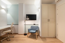 Interior Of Room With White Walls, Wardrobe, An Entrance Door And An Air Conditioner Above It. Blue Lounge Chair Bright Lights And TV On Wall, Table And Leather Office Chair For Online Remote Work.