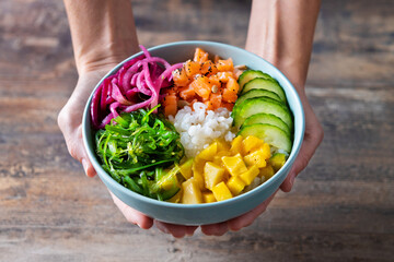 Wall Mural - Woman hands holding a poke bowl with rice, salmon,cucumber,mango,onion,wakame salad, poppy seeds ands sunflowers seeds on wooden background