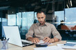 © Liubomir - Serious thinking hispanic businessman working with documents and invoices inside office, man behind paper work, using laptop at work sitting at desk.