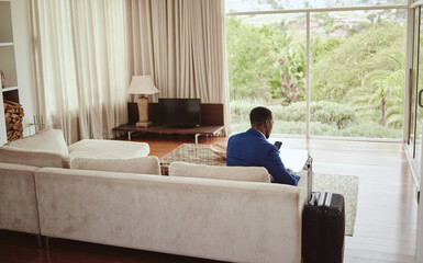 Wall Mural - Phone, travel and hospitality with a business black man sitting on a sofa in the living room of a hotel with his luggage. Back, bag and communication with a male employee typing a text message