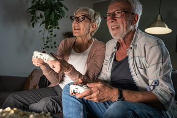 Wall Mural - Senior couple sitting on sofa and playing video game on console. They're holding a game pad and challenge each other to win.