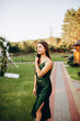 Beautiful smiling girl in a dark green dress with straps. Cute model girl stands on green grass and smiles. Brunette in the summer at a party. Emerald satin dress