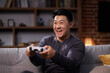 © YURII MASLAK - Happy Asian man using a wireless joystick and playing video games at home. Joyful man using PlayStation and competing in virtual games while resting on the sofa