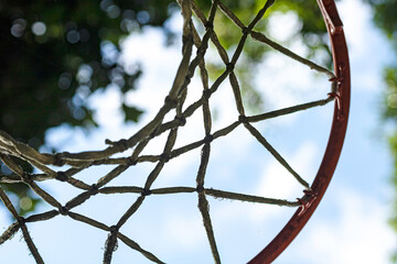 close-up de um cesta de basquete com as cordas gastos e sujos e de aro laranja e ao fundo o céu azul e algumas folhas de arvores desfocadas. 