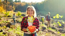 Portrait Shot Of Blonde Young Beautiful Caucasian Woman Standing Outdoor With Tree Seedling In Pot And Smiling Cheerfully. Pretty Female Eco Activist Working Against Deforestation. Gardening.