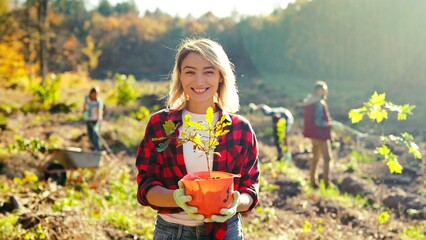 Portrait shot of blonde young beautiful Caucasian woman standing outdoor with tree seedling in pot and smiling cheerfully. Pretty female eco activist working against deforestation. Gardening.