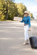 Woman With Hair Blowing In Wind Try To Stop Passing Car With Cardboard Poster On Empty Highway. Lady In Hat With Suitcase Escape From City To Go Anywhere. Travelling, Hitchhiking, Vacations, Autumn
