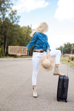 Woman With Suitcase And Cardboard Poster Stop Passing Car On Empty Highway And Look Back. Lady In Hat Escape From City To Go Anywhere. Travelling, Free Transportation, Hitchhiking, Vacations