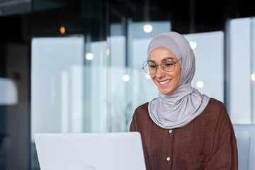 Wall Mural - Young beautiful muslim woman in hijab working inside modern office, businesswoman using laptop at work, typing on keyboard while sitting at workplace smiling and happy with achievement result.