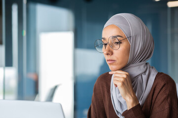 Close up of thinking business woman in hijab in modern office, serious muslim woman looking at laptop screen thinking about decision, woman at workplace successful.