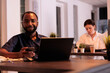 © DC Studio - Smiling african american man working on startup project on laptop, using smartphone appplication in coworking space portrait. Cheerful manager looking at camera at workplace desk
