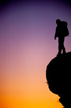 Man With A Backpack Standing On A Rock Against A Sunset In Joshua Tree NP, California (Silhouette).