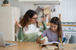 Optimistic focused teenage girl studying school curriculum reading books doing tasks under supervision of nanny. Young casual woman waving hands sits at kitchen table together with school age daughter