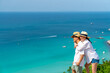 Happy Asian family couple enjoy outdoor lifestyle travel tropical island on summer holiday vacation. Husband and wife embracing each other at mountain peak and looking to blue ocean in sunny day.