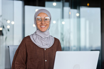 Wall Mural - Portrait of successful businesswoman inside office with laptop, woman in hijab smiling and looking at camera, muslim office worker wearing glasses.