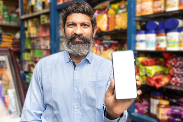 Portrait of happy mature beard Indian man showing smart phone with blank display screen to put advertisement at grocery shop or supermarket, Closeup.