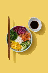 Wall Mural - Poke bowl with rice, salmon,cucumber,mango,onion,wakame salad, poppy seeds ands sunflowers seeds on yellow background. Top view