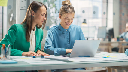 Wall Mural - Portrait of Two Creative Colleagues Collaborating and Pointing at the Laptop Screen in Office Hub. White Female Designer Discussing Work with Assistant and Choosing Marketing Strategy