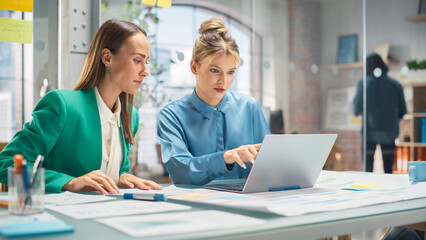 Wall Mural - Two White Females Have a Discussion in Meeting Room Behind Glass Walls in an Agency. Creative Director and Project Manager Compare Business Results and App Designs on Laptop in an Office