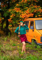 Young beautiful blonde girl with blue eyes and braided hair.Attractive model in green knitted sweater and beret standing near old retro orange bus with autumn background. Colorfull art.