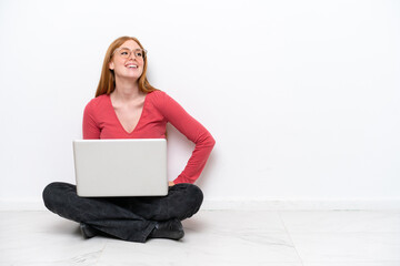 Wall Mural - Young redhead woman with a laptop sitting on the floor isolated on white background posing with arms at hip and smiling