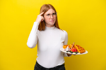 Wall Mural - Young redhead woman holding waffles isolated on yellow background having doubts and thinking