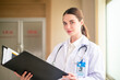 © Suriyo - Woman doctor holding folder in hospital background medicine, profession and healthcare concept - happy smiling female doctor in white coat with folder and stethoscope over hospital background