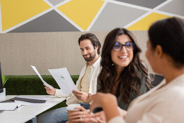 smiling manager looking at documents near cheerful interracial colleagues talking on blurred foreground.