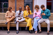 © Roman - Group of young business workers smiling happy and confident, outdoors. Posing together with smile on face using smartphones and laptop, men and women in stylish clothes sit on bench, coworking