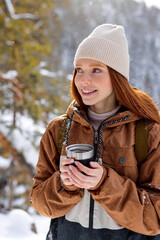 Wall Mural - female holding mug of tea. redhead lady drink from thermo mug of coffee in nature. Warm up in frozen winter forest, warm hands. travel, wanderlust, Winter hike. Outdoor recreation on frosty day.