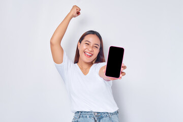 Wall Mural - Beautiful excited Asian girl wearing casual white t-shirt showing mobile phone with blank white screen, raising fist, celebrating good luck isolated on white background. People lifestyle concept