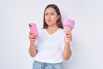 Wall Mural - Sad young blonde woman girl Asian wearing casual white t-shirt using mobile phone and holding money rupiah banknotes isolated on white background. Financial investment concept