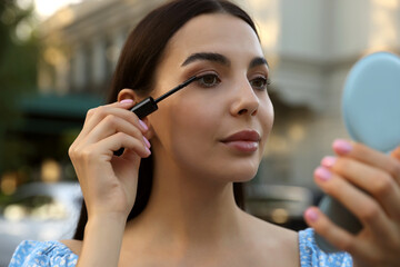 Wall Mural - Beautiful woman with cosmetic pocket mirror applying mascara outdoors, closeup