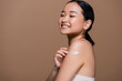 © LIGHTFIELD STUDIOS - Cheerful asian model applying cosmetic cream on shoulder isolated on brown.