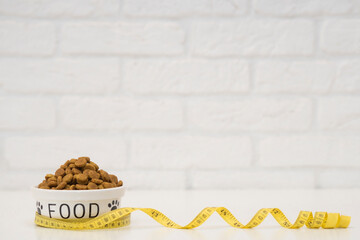 A white bowl of dry food wrapped with a yellow measuring tape against a white brick wall. The concept of a balanced diet and the fight against obesity in pets. copy space.