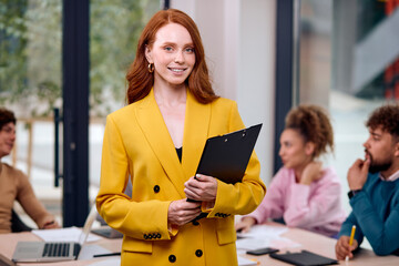 Redhead Smiling Woman With Clipboard In Hands Posing In Office During Business Meeting With Colleagues In Background, Beautiful Caucasian Lady In Stylish Yellow Suit Is Looking Confident